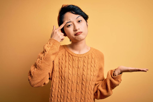 Mladá krásná asijská dívka v ležérní svetr a diadém stojící nad žlutým pozadím zmatená a otrávená s otevřenou dlaní ukazující kopírovací prostor a ukazující prstem na čelo. Přemýšlej o tom.. - Fotografie, Obrázek
