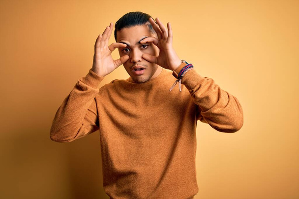 Young brazilian man wearing casual sweater standing over isolated yellow background Trying to open eyes with fingers, sleepy and tired for morning fatigue - Photo, Image