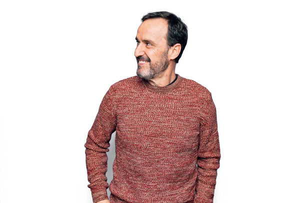 Красивый мужчина средних лет носить случайный свитер стоя на изолированном белом фоне глядя в сторону, расслабиться профиль позировать с естественным лицом и уверенной улыбкой
. - Фото, изображение