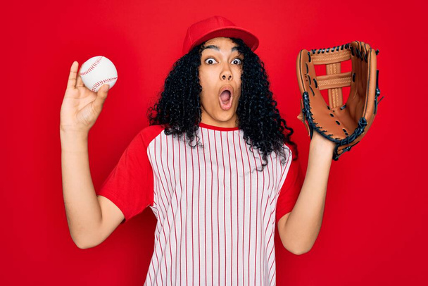 Young african american curly sportswoman wearing cap playing baseball using ball and glove scared in shock with a surprise face, afraid and excited with fear expression - Fotoğraf, Görsel