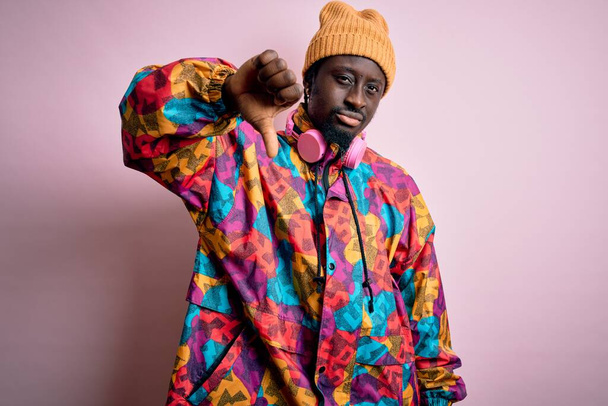 Young handsome african american man wearing colorful coat and cap over pink background looking unhappy and angry showing rejection and negative with thumbs down gesture. Bad expression. - Photo, Image