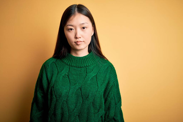 Jeune belle femme asiatique portant un pull d'hiver vert sur fond jaune isolé Détendu avec une expression sérieuse sur le visage. Un regard simple et naturel sur la caméra
. - Photo, image