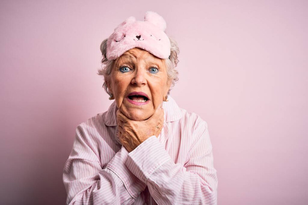Senior mooie vrouw met slaapmasker en pyjama over geïsoleerde roze achtergrond schreeuwen en stikken omdat pijnlijke wurging. Gezondheidsproblemen. Verstikking en zelfmoordconcept. - Foto, afbeelding