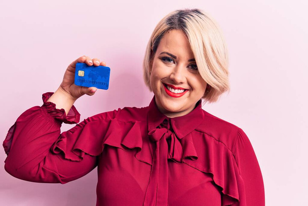 Young beautiful blonde plus size woman holding credit card over isolated pink background looking positive and happy standing and smiling with a confident smile showing teeth - Photo, Image