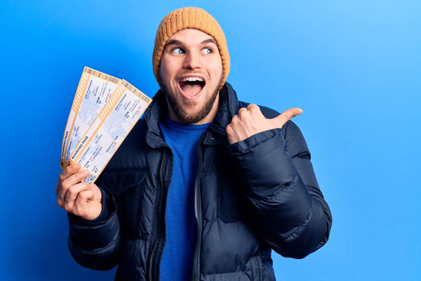Young handsome man wearing winter clothes holding airline boarding pass pointing thumb up to the side smiling happy with open mouth  - Foto, imagen