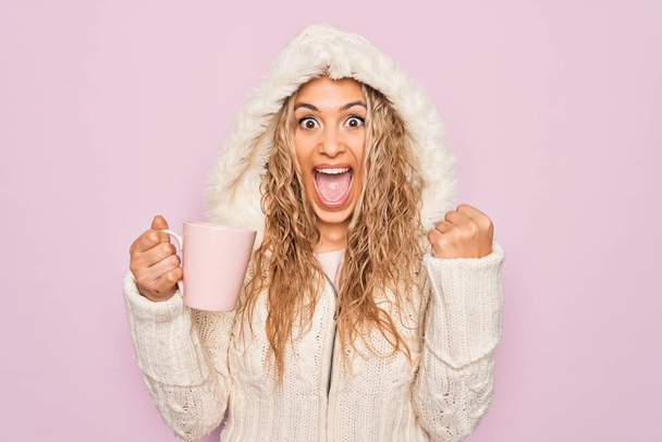 Junge schöne blonde Frau in lässigem Pullover mit Kapuze trinkt Tasse Kaffee schreit stolz, feiert Sieg und Erfolg sehr aufgeregt mit erhobenen Armen - Foto, Bild