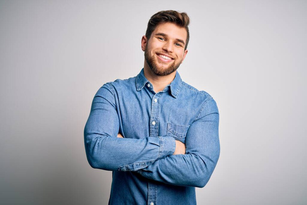 Young handsome blond man with beard and blue eyes wearing casual denim shirt happy face smiling with crossed arms looking at the camera. Positive person. - Photo, Image