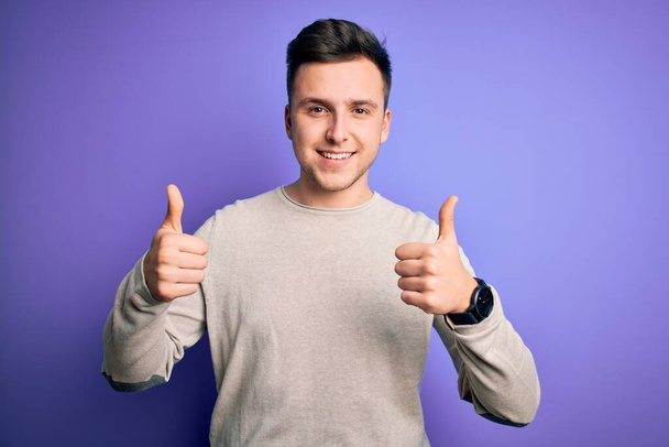Молодой красивый белый мужчина в случайном свитере на фиолетовом изолированном фоне знак успеха делает позитивный жест с рукой, большие пальцы вверх улыбается и счастлив. Веселое выражение лица и жест победителя
. - Фото, изображение