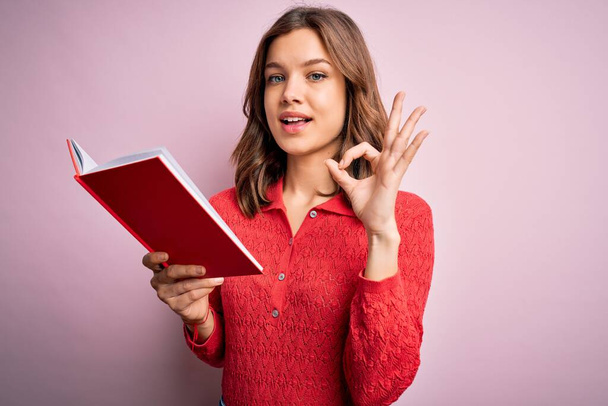Fiatal szőke diák lány olvas egy könyvet rózsaszín elszigetelt háttér csinál rendben jel ujjakkal, kiváló szimbólum - Fotó, kép