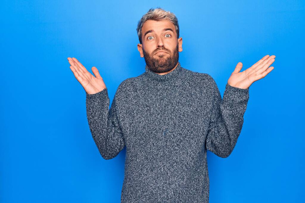 Young handsome blond man wearing casual sweater standing over isolated blue background clueless and confused with open arms, no idea and doubtful face. - Photo, Image