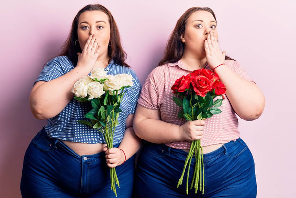 Молодые близнецы, держащие цветы, покрывающие рот рукой, шокированы и боятся ошибиться. Удивительное выражение  - Фото, изображение