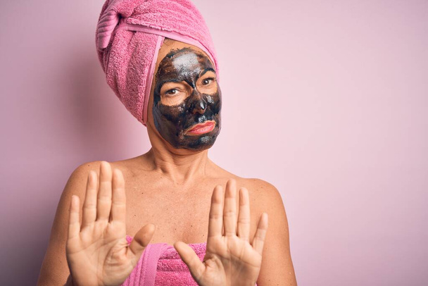 Брюнетка средних лет носит красивую черную маску на изолированном розовом фоне, отталкивая ладони, показывая отказ и отрицание со страшным и отвратительным выражением лица. Остановить и запретить
. - Фото, изображение