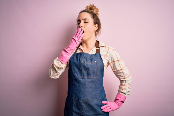 Young beautiful blonde cleaner woman doing housework wearing arpon and gloves bored yawning tired covering mouth with hand. Restless and sleepiness. - Photo, Image
