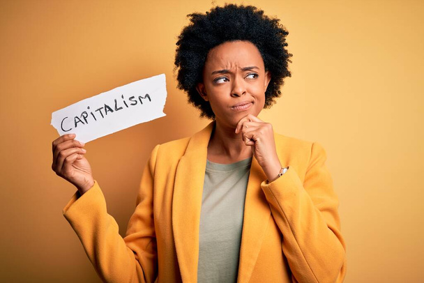 African American afro businesswoman with curly hair holding paper with capitalism message serious face thinking about question, very confused idea - Photo, Image