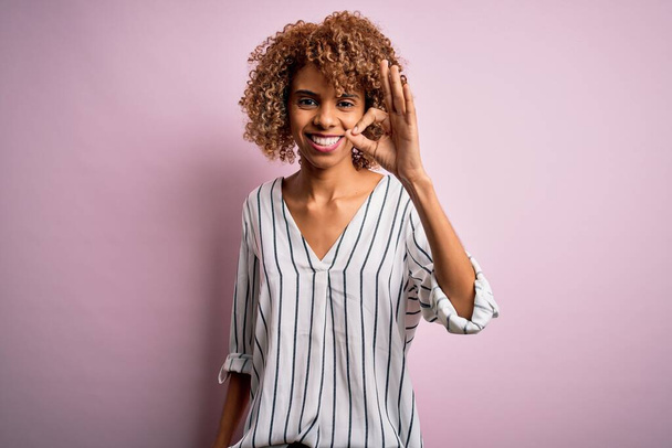 Beautiful african american woman with curly hair wearing striped t-shirt over pink background smiling positive doing ok sign with hand and fingers. Successful expression. - Foto, Imagem