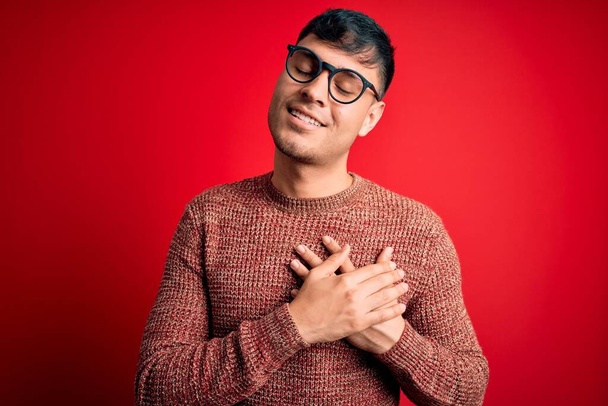 Homem hispânico bonito jovem usando óculos nerd sobre fundo vermelho sorrindo com as mãos no peito com os olhos fechados e gesto grato no rosto. Conceito de saúde
. - Foto, Imagem