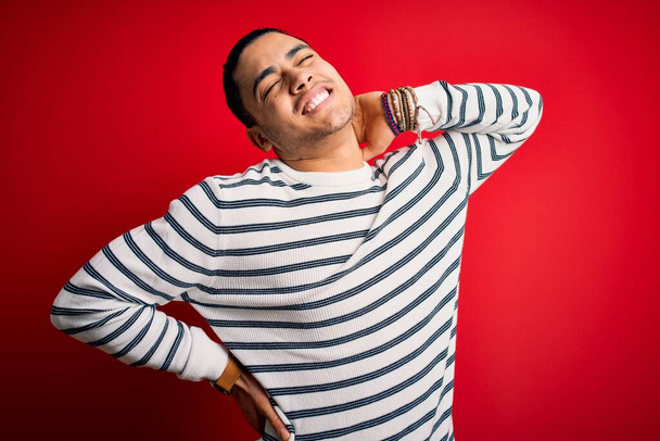 Young brazilian man wearing casual striped t-shirt standing over isolated red background Suffering of neck ache injury, touching neck with hand, muscular pain - Foto, Imagen