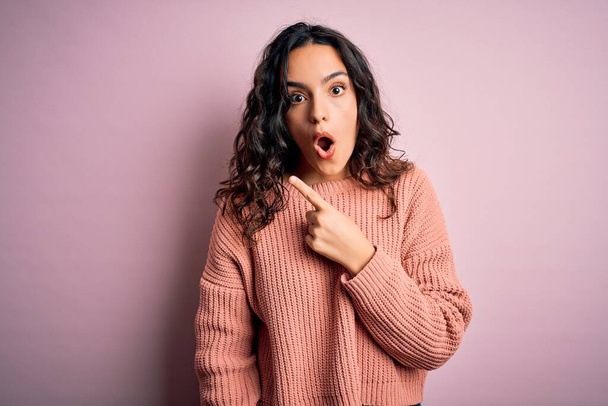 Young beautiful woman with curly hair wearing casual sweater over isolated pink background Surprised pointing with finger to the side, open mouth amazed expression. - Foto, Imagem