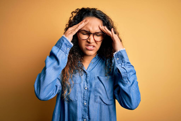 Beautiful woman with curly hair wearing casual denim shirt and glasses over yellow background with hand on head for pain in head because stress. Suffering migraine. - Фото, зображення
