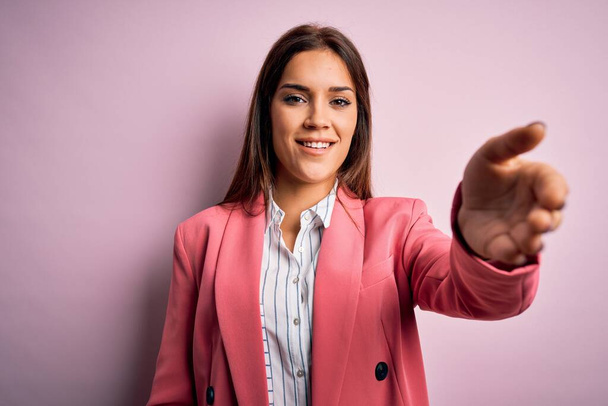 Jonge mooie brunette vrouw dragen jas staande over geïsoleerde roze achtergrond glimlachend vriendelijk aanbieden van handdruk als groet en gastvrij. Succesvolle bedrijfsvoering. - Foto, afbeelding