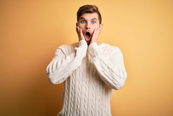 Young blond man with beard and blue eyes wearing white sweater over yellow background afraid and shocked, surprise and amazed expression with hands on face - 写真・画像