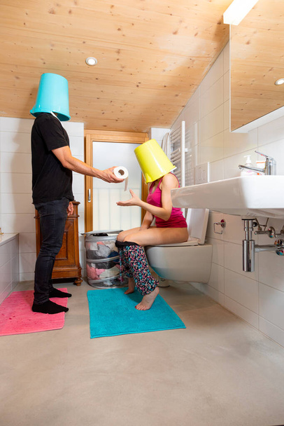 Man with bucket on his head passes the toilet paper to his wife sitting on the toilet, she too with bucket on her head. Funny situation. - Zdjęcie, obraz