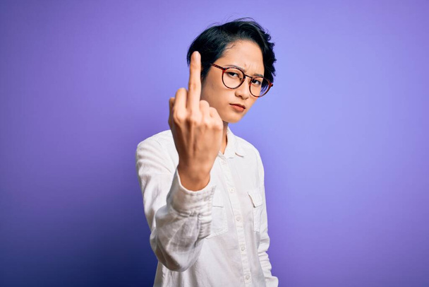 Young beautiful asian girl wearing casual shirt and glasses standing over purple background Showing middle finger, impolite and rude fuck off expression - Φωτογραφία, εικόνα