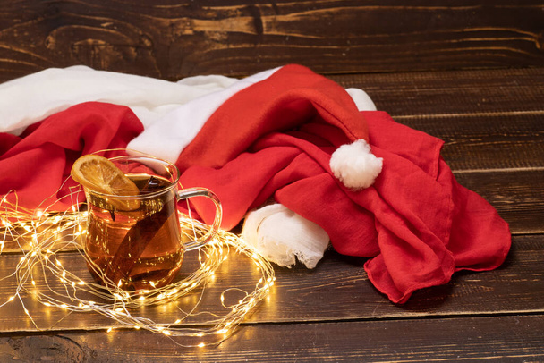 クリスマスのコンセプト。サンタの帽子、照明付きのガーランド、暗い木製の背景に熱い飲み物のカップで構成されています。冬のお茶、シナモン、オレンジのスライス。休日の香り。反射だ。スペースのコピー. - 写真・画像