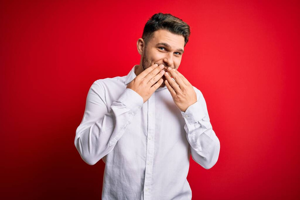 Young business man with blue eyes wearing elegant shirt standing over red isolated background laughing and embarrassed giggle covering mouth with hands, gossip and scandal concept - Photo, Image