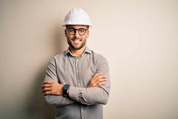 Young architect man wearing builder safety helmet over isolated background happy face smiling with crossed arms looking at the camera. Positive person. - Fotoğraf, Görsel