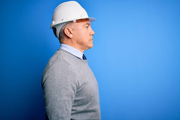Ingeniero guapo de pelo gris de mediana edad que usa casco de seguridad sobre fondo azul mirando hacia un lado, pose de perfil relajado con cara natural con sonrisa confiada
. - Foto, imagen