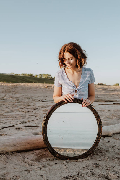 Outdoor foto stile di vita atmosferico di giovane bella donna dai capelli scuri in abito blu seduto sulla spiaggia e tenendo specchio.Foto concettuale con uno specchio
. - Foto, immagini