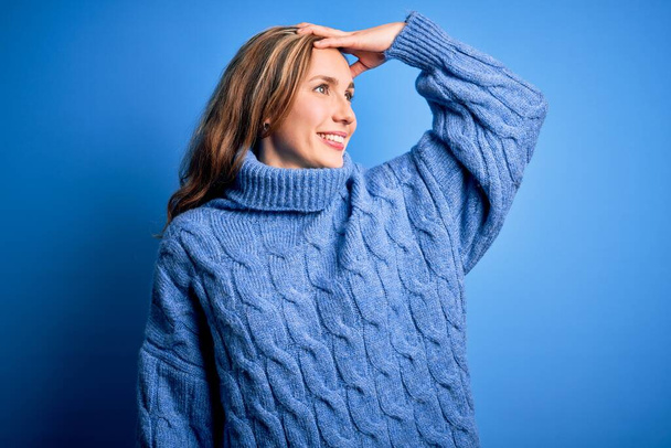Joven hermosa mujer rubia con suéter casual cuello alto sobre fondo azul sonriendo seguro de tocar el cabello con la mano hacia arriba gesto, posando atractivo y de moda
 - Foto, imagen