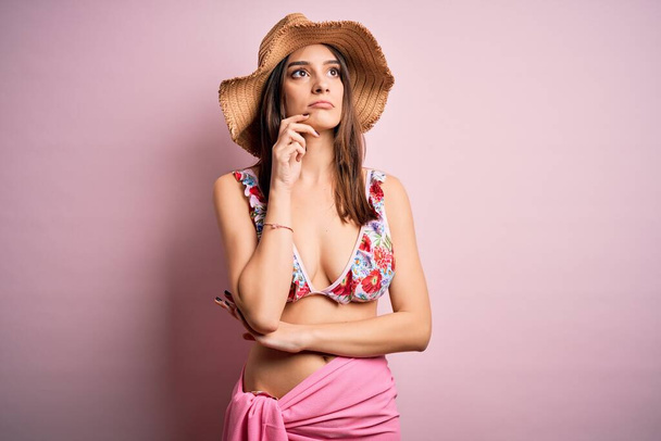 Молодая красивая женщина в отпуске в бикини и летняя шляпа на розовом фоне с рукой на подбородке думает о вопросе, задумчивое выражение лица. Улыбается с вдумчивым лицом. Концепция сомнений
. - Фото, изображение