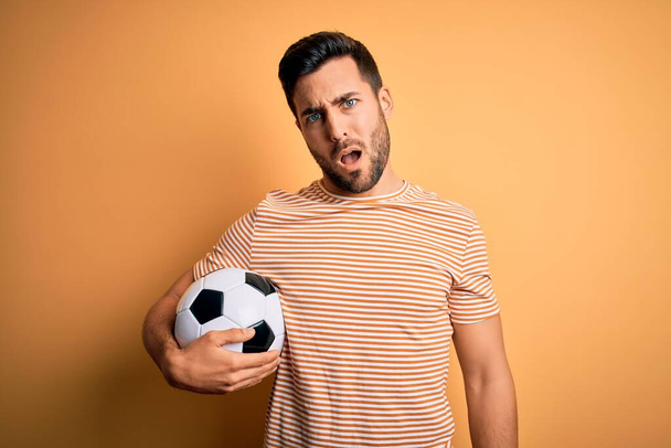 Όμορφος παίκτης με γενειάδα που παίζει ποδόσφαιρο κρατώντας μπάλα πάνω από κίτρινο φόντο στο πρόσωπο σοκ, δείχνοντας σκεπτικός και σαρκαστικός, έκπληκτος με ανοιχτό στόμα - Φωτογραφία, εικόνα