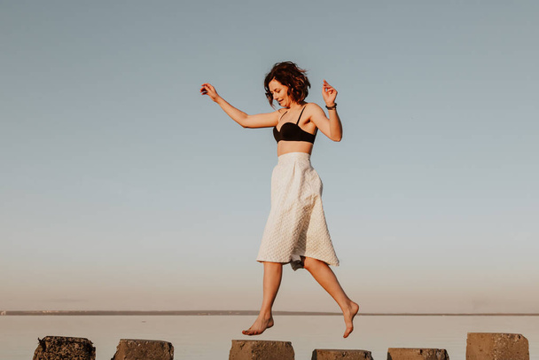 Posing woman in nature. Young woman in stylish clothes:  top and white skirt  jumping on stones  against the backdrop of a  sea. Photos on summer vacation  - Foto, Bild
