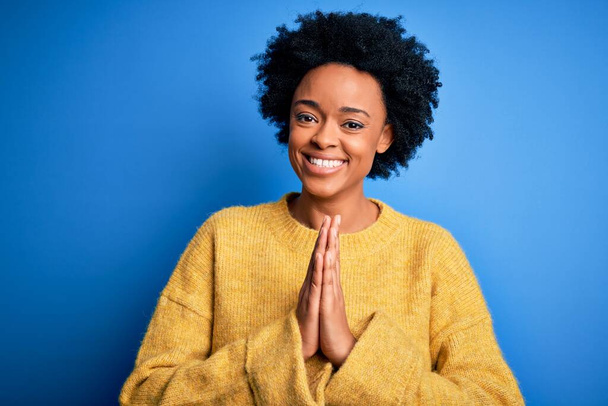 Junge schöne afroamerikanische Afro-Frau mit lockigem Haar trägt einen gelben lässigen Pullover und betet mit den Händen zusammen und bittet selbstbewusst um Vergebung. - Foto, Bild