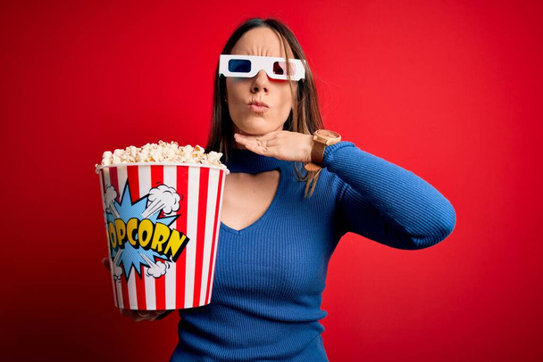 Νεαρή ξανθιά γυναίκα που φοράει 3d γυαλιά και τρώει ποπ κορν βλέποντας μια ταινία στον κινηματογράφο να κόβει το λαιμό με το χέρι ως μαχαίρι, απειλεί την επιθετικότητα με έξαλλη βία - Φωτογραφία, εικόνα