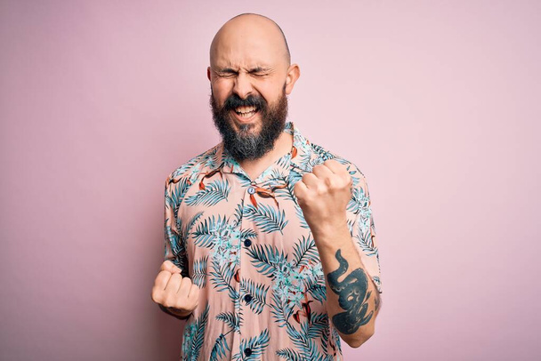 Handsome bald man with beard and tattoo wearing casual floral shirt over pink background celebrating surprised and amazed for success with arms raised and eyes closed. Winner concept. - Photo, Image