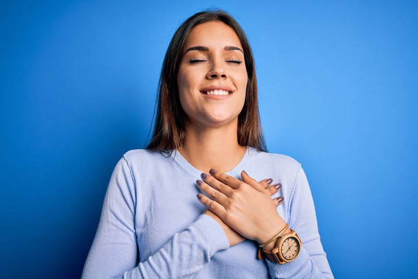 Young beautiful brunette woman wearing casual sweater standing over blue background smiling with hands on chest with closed eyes and grateful gesture on face. Health concept. - Foto, immagini