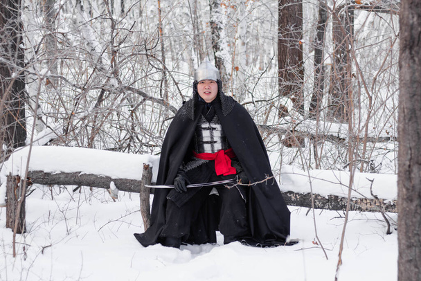   鎖状の郵便鎧を着た中世の戦士、ヘルメットと彼の手にサーベルと黒のマントは、木に脇に立っています。冬の森や雪を背景に. - 写真・画像