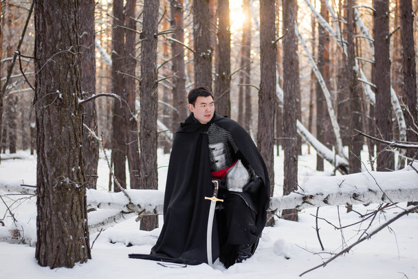   A medieval warrior in chain mail armor, a helmet and a black cloak with a saber in his hands, stands aside to a tree. Against the background of winter forest and snow. - 写真・画像