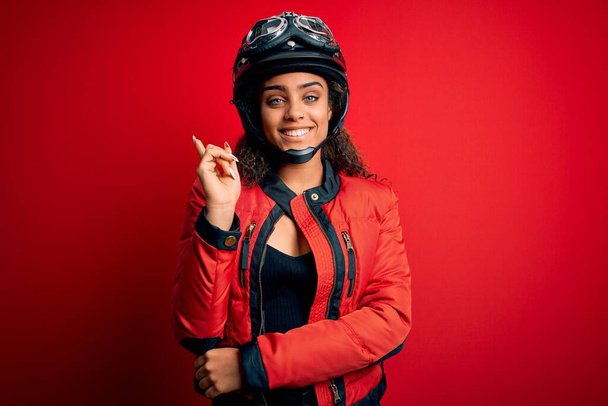 Young african american motorcyclist girl wearing moto helmet and glasses over red background with a big smile on face, pointing with hand and finger to the side looking at the camera. - Фото, изображение