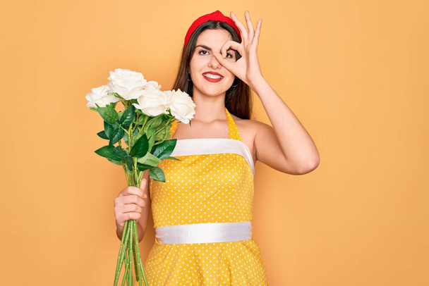 Νεαρή όμορφη pin up γυναίκα φορώντας 50 μόδας vintage φόρεμα κρατώντας boquet τριαντάφυλλα λουλούδια με χαρούμενο πρόσωπο χαμογελώντας κάνει ok σημάδι με το χέρι στο μάτι κοιτάζοντας μέσα από τα δάχτυλα - Φωτογραφία, εικόνα