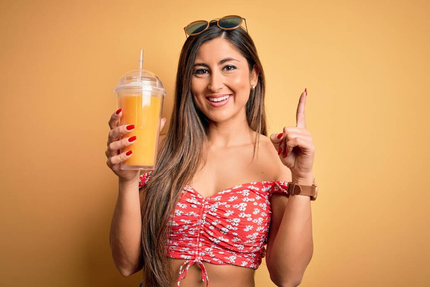 Piękna brunetka pijąca szklankę ze zdrowym sokiem pomarańczowym na żółtym tle zaskoczona pomysłem lub pytaniem wskazującym palcem na szczęśliwą twarz, numer jeden - Zdjęcie, obraz