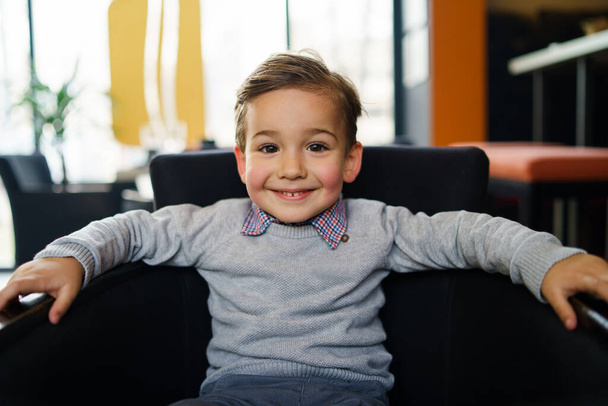 Porträt eines kleinen dreijährigen kaukasischen Jungen, der auf dem Stuhl sitzt und lächelnd in die Kamera blickt - Foto, Bild