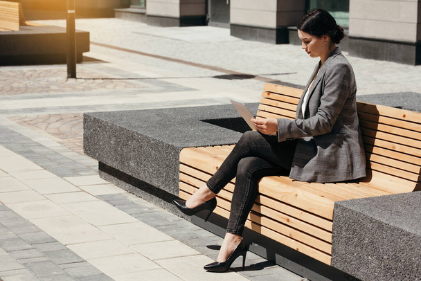 Attractive girl in business clothes sits on a bench in the city center and looks at the news on a laptop - Foto, Imagem