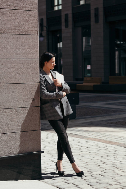 Vertical photo of businesswoman in plaid coat and trousers leaning on the corner of a building and holding a laptop in her hands. - Photo, Image