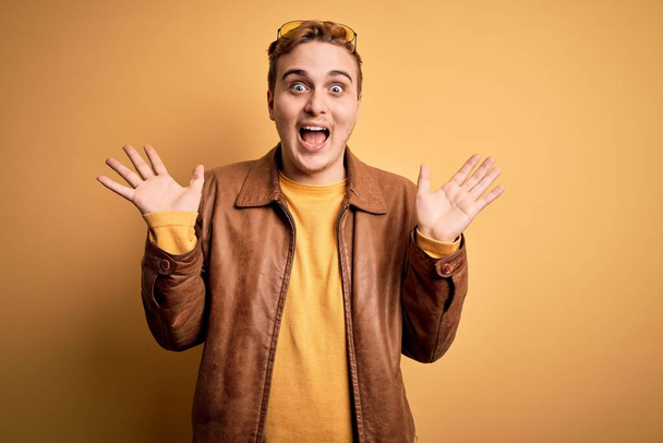 Jeune homme rousse beau portant une veste en cuir décontractée sur fond jaune isolé célébrant fou et étonné pour le succès avec les bras levés et les yeux ouverts hurlant excités. Concept gagnant
 - Photo, image