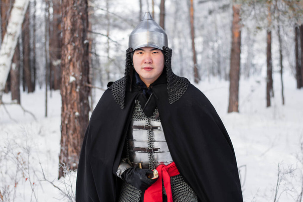   Portrait of a medieval warrior in armor, a helmet and a black cloak with a saber in his hands against the background of a winter forest and snow. - 写真・画像